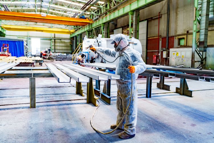 Worker in protective clothing spray-coating metal beams inside an industrial factory.