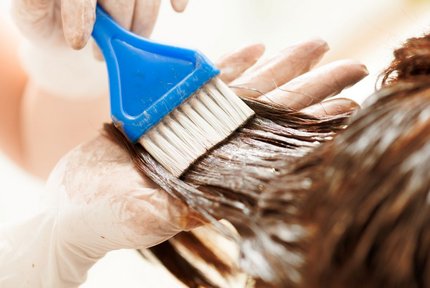 Gloved hands using a blue applicator brush to apply brown hair dye close-up.
