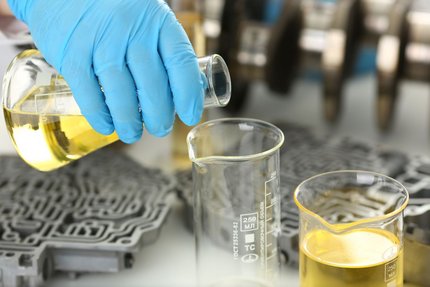 Gloved hand pouring yellow liquid into a beaker in an industrial lab setting.
