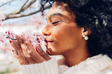 Close-up of a woman with eyes closed, smelling pink cherry blossoms on a branch outdoors.