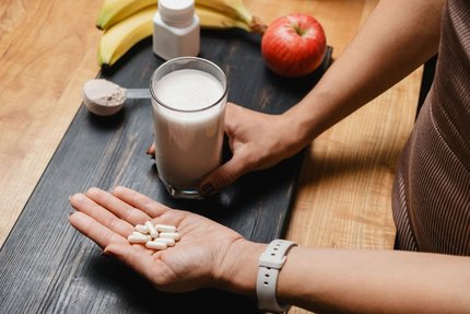 Hand holding supplement capsules beside a milk shake, protein powder, and fresh fruit.