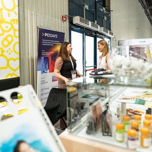 Two women talk beside a glass display of cosmetic samples at HSH Chemie’s booth during PCI Days 2026.