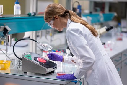 Scientist in safety glasses and gloves examines a sample in a Petri dish at a laboratory workstation.