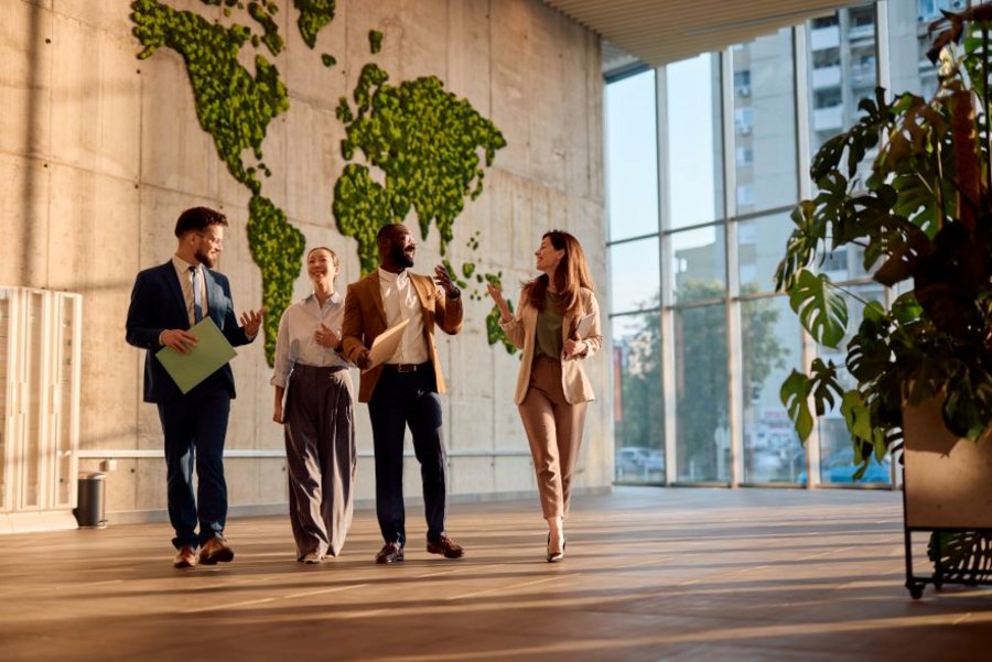Four business professionals walking in a modern office lobby with a green world map wall and large windows.