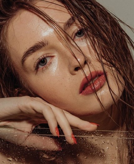 Close-up portrait of a woman's dewy, glowing skin and wet hair near a water-droplet surface.