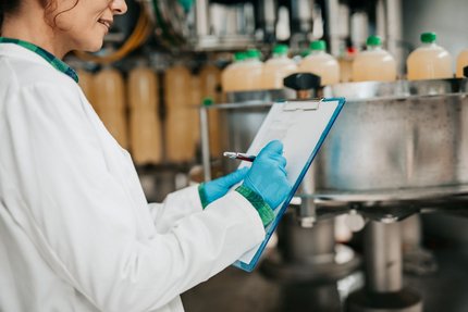 Scientist in a lab coat and blue gloves records observations on a clipboard beside industrial processing equipment.