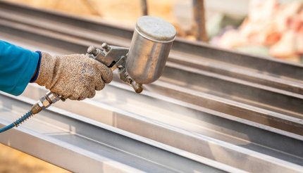 Gloved worker uses a spray gun to apply a coating to long metal beams in an industrial workshop.
