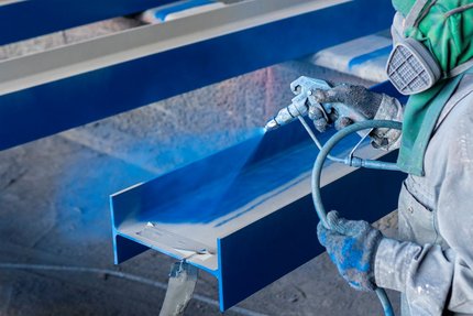 Worker in protective gear spray-paints a blue coating onto a steel beam in an industrial workshop.